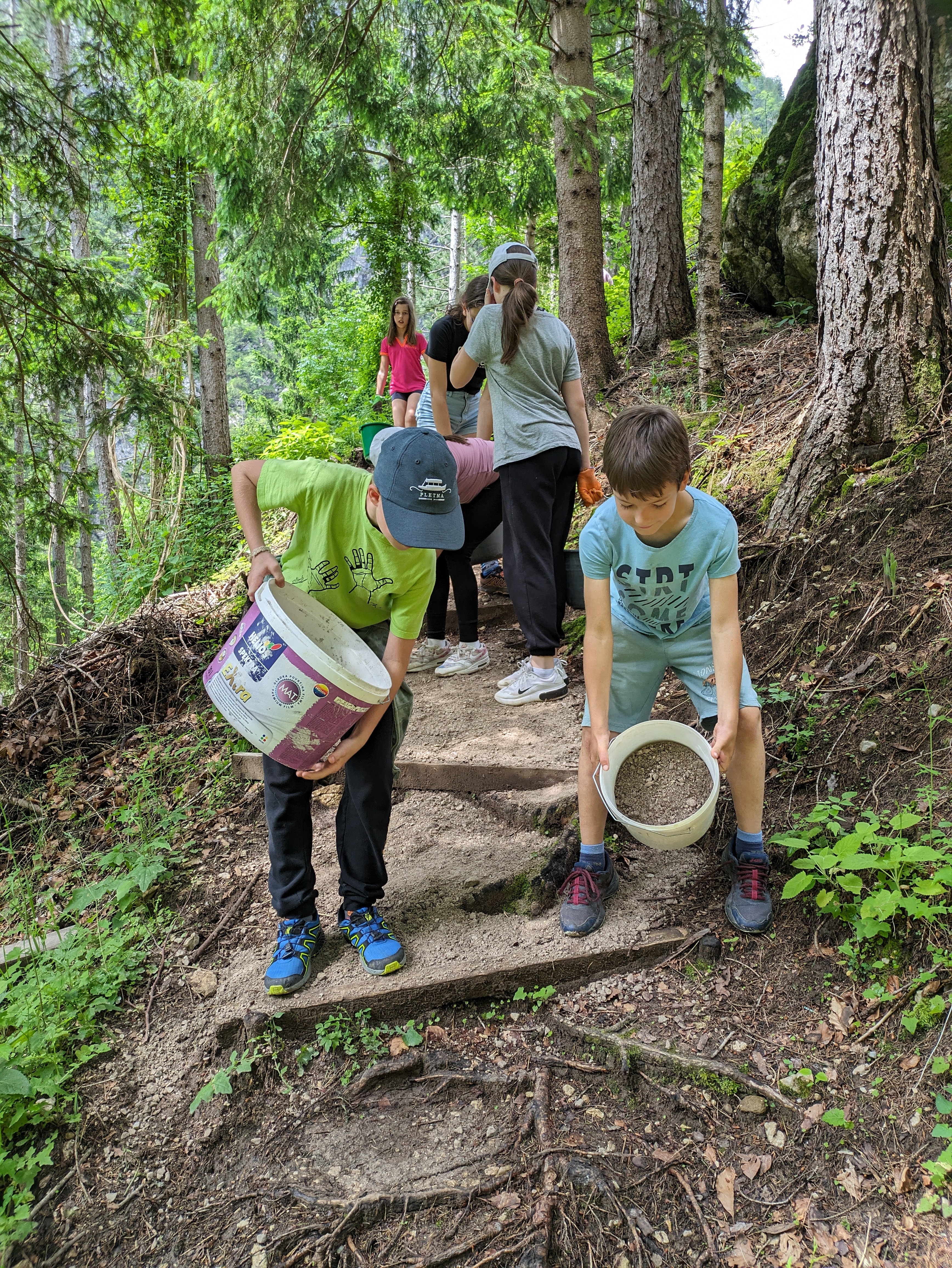 Na fotografiji so otroci, ki v roki nosijo veder napolnjen s fino zemljo in jo sipajo po gozdni stezi. Okoli njih je smrekov gozd.
